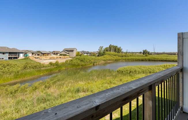 A wooden deck overlooks a grassy field with a river and houses in the distance.