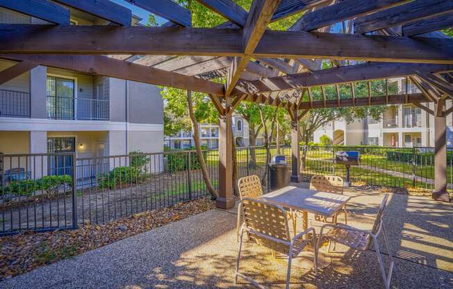 Outdoor seating area with a wooden pergola at Saxony at Chase Oaks Apartments in Dallas, TX, featuring a table and chairs, surrounded by lush landscaping and views of apartment balconies.