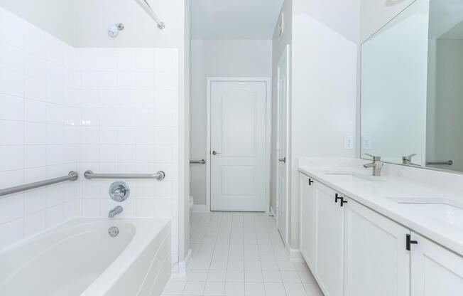 A well-lit bathroom featuring a white tiled floor, a bathtub with a grab bar, a large mirror above a double sink with modern fixtures, white cabinetry, and a plain white door leading to another room, all set against a bright, neutral wall.