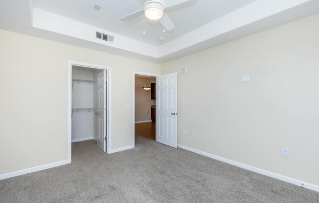 A well-lit room with beige walls and light brown carpet. There are two doors: one leading to a closet and the other to a hallway, with a ceiling fan overhead. The room features modern design elements and natural light, creating a spacious feel.