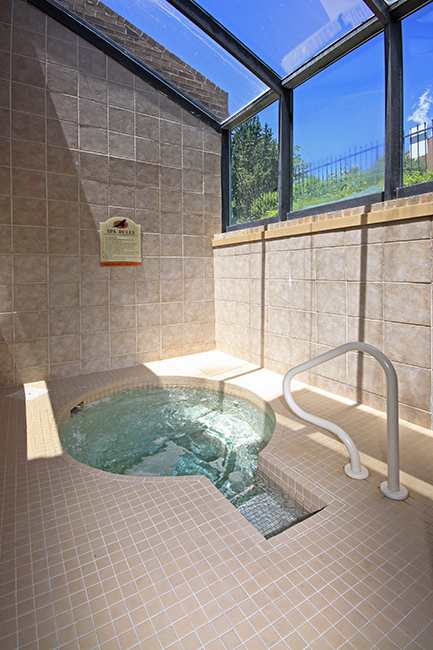 A bright indoor spa area here at Eagle Ridge featuring a tiled hot tub with gently bubbling water, a built-in handrail for easy access, neutral tile walls and flooring, and large angled windows overhead that allow abundant natural light and views of greenery outside.