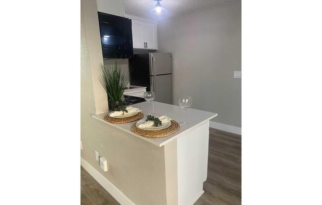A modern kitchen space featuring a white countertop with two decorative plates topped with plants, alongside two wine glasses. The kitchen has black cabinetry, stainless steel appliances, and a stylish light fixture above. The flooring is a light wood color, creating a bright and inviting atmosphere.