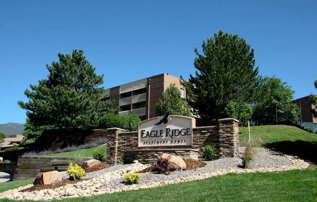 A welcoming stone monument sign here at Eagle Ridge set on a gently sloped landscaped hillside, featuring layered stonework, gravel accents, and low shrubs, with mature pine trees and a multi-story apartment building rising behind it under a clear blue sky.