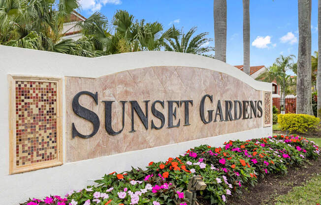 Stone monument sign placed reads Sunset Gardens, located outdoors in a flower bed near the entrance driveway