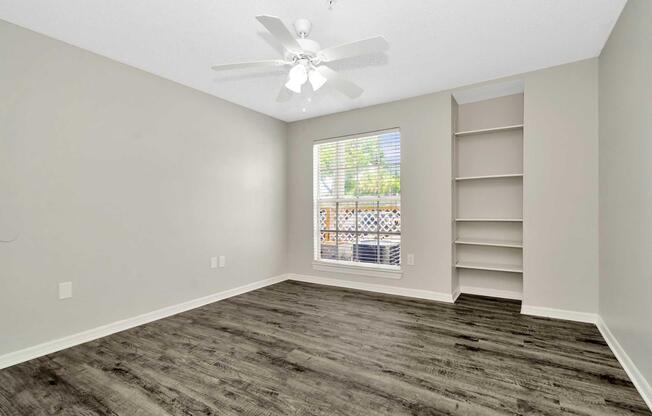A well-lit, empty bedroom featuring light gray walls, a ceiling fan, and a window with blinds. The floor is dark wood, and there's a small built-in shelf next to the window. Natural light floods the space, emphasizing its clean and spacious feel.