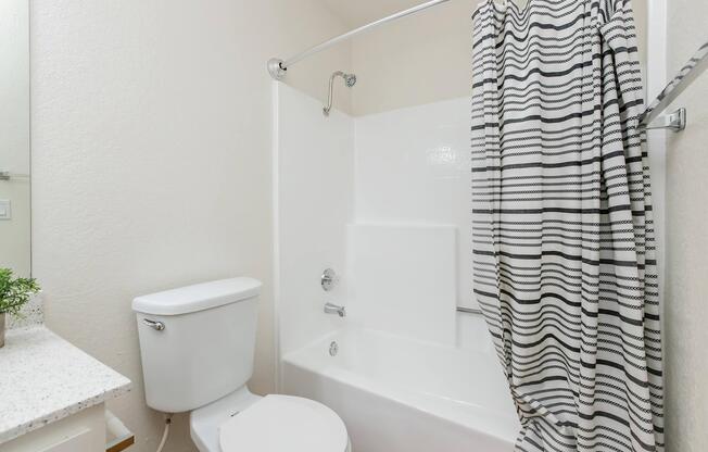 A clean bathroom featuring a white bathtub with a shower curtain in a striped pattern. There is a modern toilet nearby and a countertop with a small potted plant. The walls are painted in a light color, creating a bright and airy atmosphere.