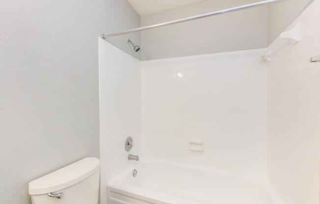 A clean, well-lit bathroom with a white bathtub and shower, featuring a silver showerhead. The walls are painted light gray, and there's a toilet next to the bathtub. The space is uncluttered and functional, ideal for daily use.