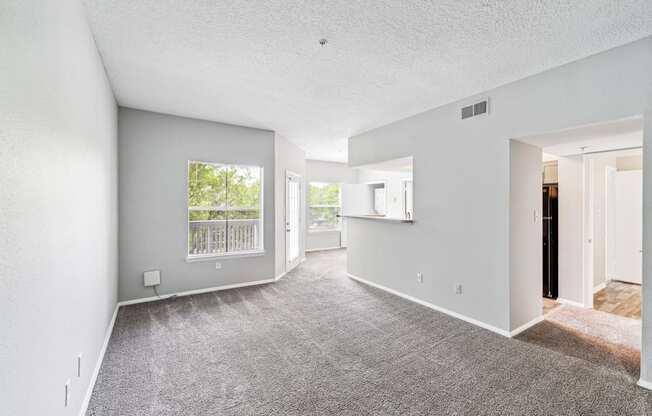 the living room and kitchen of an apartment with carpeting and a large window