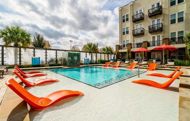 A resort-style pool here at BRIO at Firewheel with a shallow sun shelf, bright orange in-water lounge chairs, clear swimming area, poolside seating, and umbrellas beside modern apartment buildings.