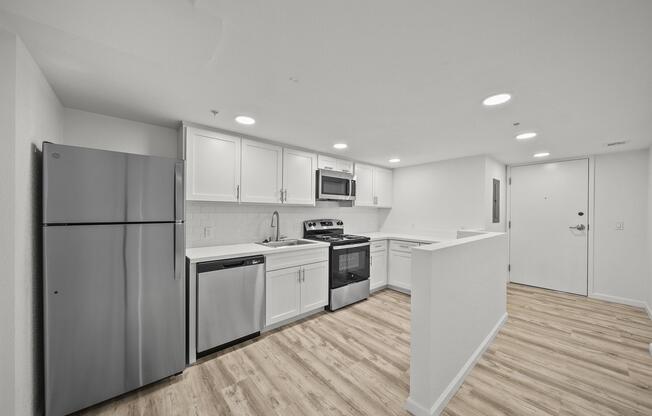 Modern kitchen featuring stainless steel appliances including a refrigerator, dishwasher, microwave, and oven. The space has light-colored cabinets and a spacious countertop with a wood-like flooring. Bright overhead lights illuminate the room, creating a welcoming atmosphere.