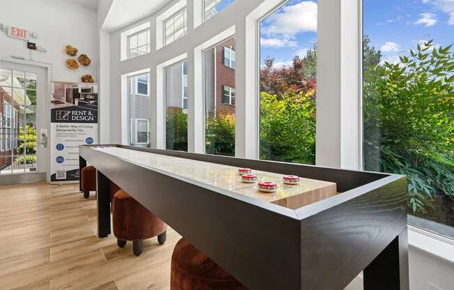 A long table with a shelf underneath it is in the foreground of a room with a window showing the outside.