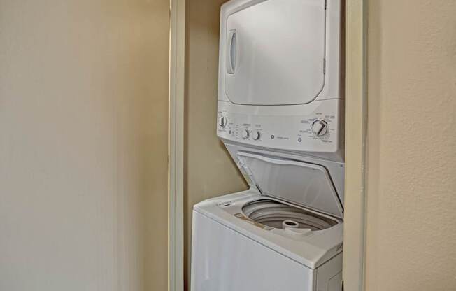 A convenient in-home laundry area here at Whispering Cedars with a stacked white washer and dryer tucked into a hallway closet, offering a practical and space-saving feature for easy everyday living.