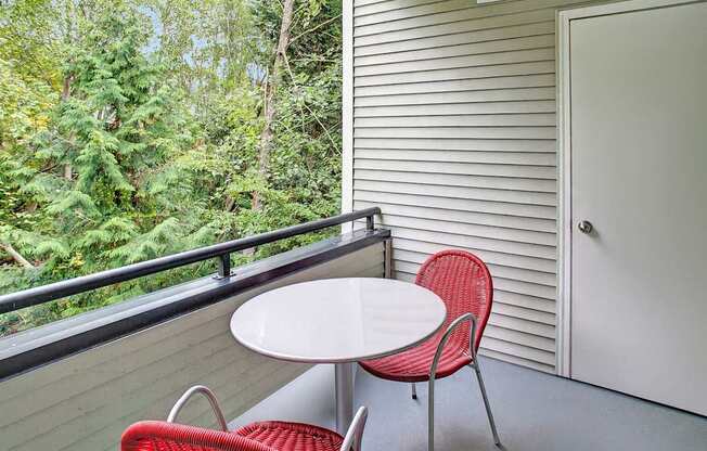 A private balcony here at Portsmith featuring a small round café table with two red woven chairs, a gray-painted floor, and a black metal railing overlooking lush green trees, with light gray exterior siding and a storage door creating a quiet outdoor space for relaxing or dining.