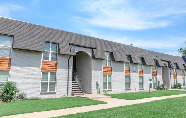 A modern multi-unit apartment building featuring a gray exterior with wooden accents, multiple windows, and a staircase leading to an upper level. The surrounding area includes neatly manicured grass and landscaping under a bright, blue sky.