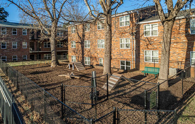 a fenced in area with a bench and trees in front of a brick building