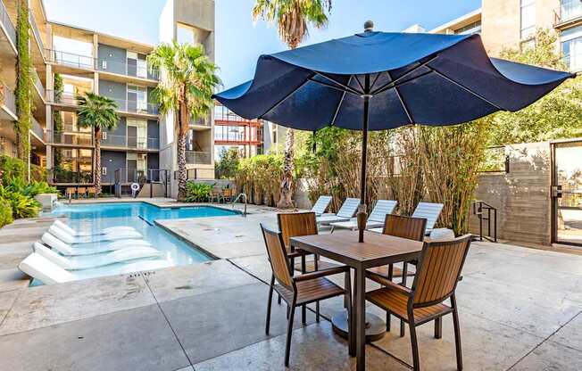 A table with chairs and an umbrella is in the foreground of a pool area.
