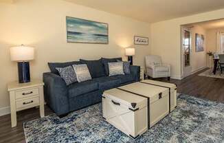 A living room with a grey couch, a white ottoman, and a painting on the wall.