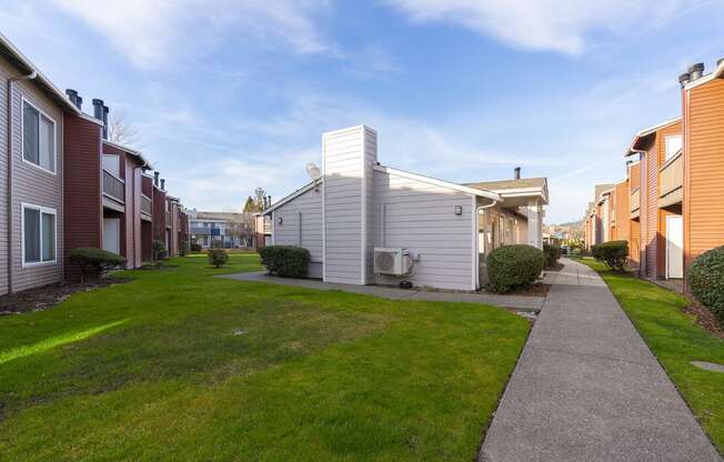 A paved pathway surrounded by green grass leads to apartments on a sunny blue sky day.