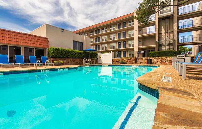 A pool area with lounge chairs, a patio table and chairs with a blue umbrella and stainless steel BBQ in front of apartment buildings at Princeton Court Apartments in the Vickery Midtown neighborhood of Dallas, TX.