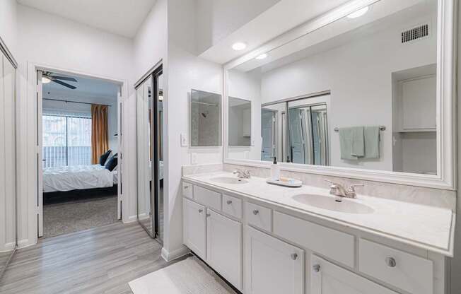 A white bathroom with a large mirror and a bedroom visible through the door.