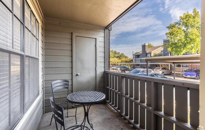 A small balcony featuring a round black metal table with two chairs, a door leading inside, and a view of parked cars and greenery outside. The walls are painted in a light tone with large windows allowing natural light. The sky is partly cloudy, creating a serene atmosphere.