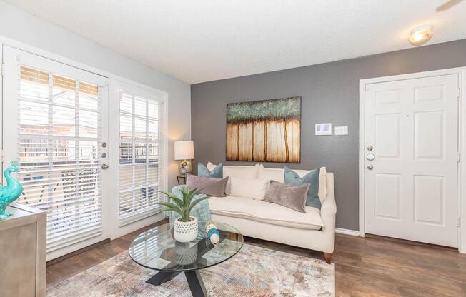 A cozy living room featuring a light-colored sofa with decorative pillows, a round glass coffee table, and a potted plant. The walls are painted gray, and a large piece of abstract art hangs above the sofa. Natural light streams through white shutters on the windows, and a welcoming entrance door is visible.