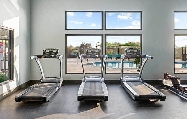 Three treadmills are lined up in a gym with a view of the outdoors through the windows.