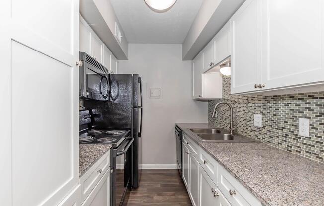 A modern kitchen featuring white cabinets, granite countertops, and a backsplash with mosaic tiles. It includes a black refrigerator, a stove, and a double sink, with natural light coming from a ceiling fixture. The floor is dark wood, creating a contemporary and inviting space.