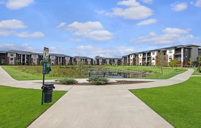 A serene view of modern 49 West Apartment buildings surrounding a central pond under a blue sky with clouds. Green lawns and pathways create a welcoming atmosphere.