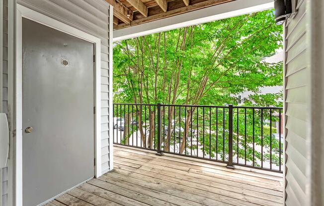A wooden balcony with a railing overlooks a lush, green tree outside. There is a plain gray door on one side, and the surrounding area is well-lit by natural sunlight, creating a serene and inviting atmosphere.