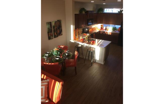 A well-lit modern kitchen and dining area featuring a dining table set for four with green tableware, a cozy living space with an orange sofa, and wooden cabinets in the kitchen. Natural light filters through the windows, highlighting potted plants and decor on the countertops.