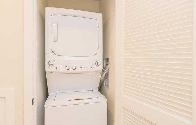 A stacked washer and dryer unit situated in a small laundry space, with a light-colored wall and a louvered door nearby. The appliance features control knobs and a front-loading washing machine on the bottom.