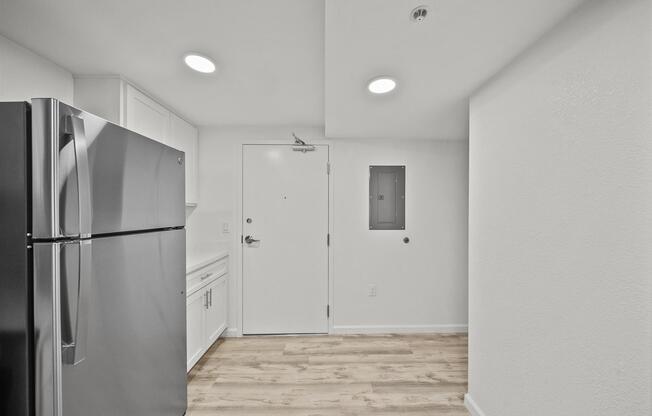 A modern kitchen area featuring a stainless steel refrigerator, white cabinets, and light-colored walls. The space includes a door leading outside, a small electrical panel on the wall, and hardwood-style flooring, creating a clean and bright atmosphere.