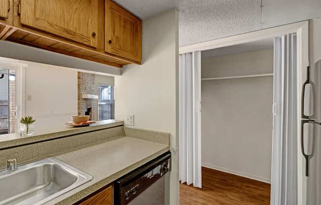 A practical kitchen and storage area here at Summertree Place featuring warm wood cabinetry above the counters, a stainless sink and dishwasher, durable laminate countertops, and an open pantry or storage nook with sliding doors that adds convenient extra space just off the kitchen.