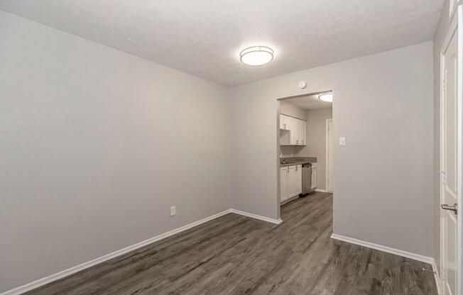 A spacious, empty room with light gray walls and a textured ceiling. The floor is finished with a contemporary laminate. Two round ceiling lights illuminate the space. An open doorway leads to a kitchen area with white cabinets in the background, enhancing the sense of openness and potential for furnishing.