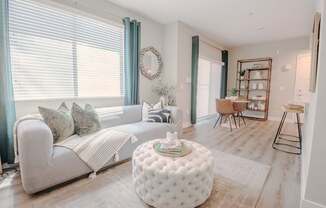 An open concept living and dining space with a white couch and a round ottoman, bar stools, dining table and bookshelf.