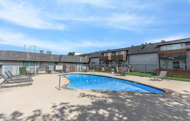 A swimming pool surrounded by lounge chairs and a fence. In the background, there are two-story residential buildings with balconies. The sky is clear with a few clouds, and greenery is visible around the pool area.