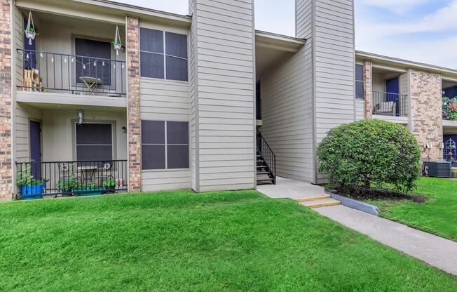 A view of a multi-unit apartment building featuring beige siding, a small set of stairs leading to the entrance, and well-maintained green grass and bushes in the foreground.
