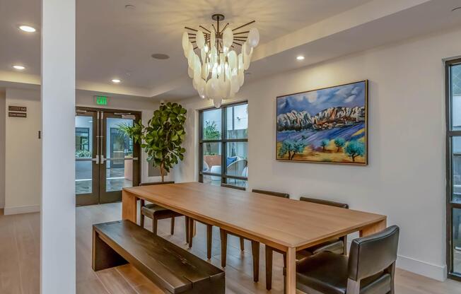 A modern interior of a dining area featuring a large wooden table with black chairs, a statement chandelier hanging from the ceiling, and a vibrant painting of a landscape on the wall. There are large windows allowing natural light and a green plant adding a touch of nature to the space.