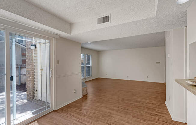 A bright open living room here at Summertree Place featuring wood-style flooring, a sliding glass door opening to a private patio, neutral walls, a textured ceiling, large windows for natural light, and an open layout that flows seamlessly toward the kitchen area, creating a spacious and inviting living space.