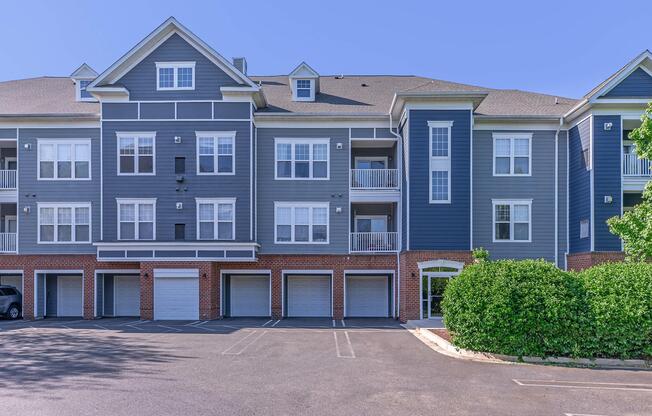 A modern apartment complex featuring a blue exterior with white trim, multiple balconies, and garage spaces on the ground level. The parking area is visible in front, with a well-maintained landscape including greenery. The sky is clear and blue.