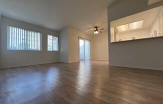 A spacious, empty living room with light-colored wooden flooring. Large windows with horizontal blinds allow natural light to fill the space. A ceiling fan provides ventilation. There's an open view to a kitchen area beyond a pass-through counter, and a sliding glass door leads to an outdoor patio.