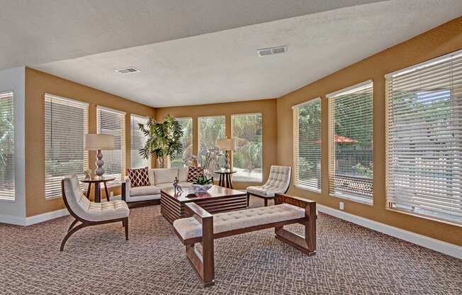 A bright community lounge here at Northridge Court featuring expansive windows with white blinds, soft neutral walls, patterned carpet flooring, a cozy seating arrangement with sofas, accent chairs, table lamps, and lush greenery that create a welcoming space for relaxing or socializing.