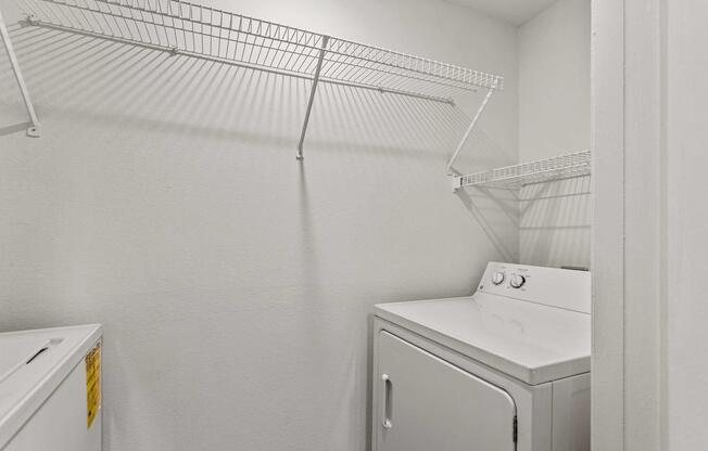 A clean, small laundry room featuring a white washer and a dryer side by side. Above, a wire shelf is mounted on a plain white wall, providing storage space. The room is well-lit and organized, emphasizing a tidy and functional laundry area.