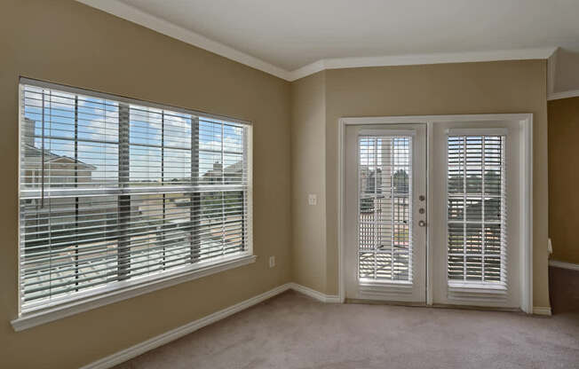 A bright and comfortable living room here at Mission Green showcasing neutral-toned walls, soft carpeted flooring, a wide window with blinds that brings in natural light, and glass patio doors with matching blinds that open to an outdoor area, creating an airy and inviting atmosphere for everyday living.
