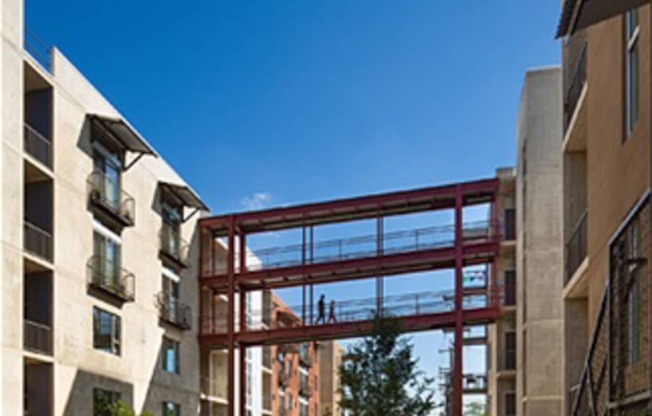 A man rides his bicycle down a sidewalk in front of a building with a large red structure in the background.