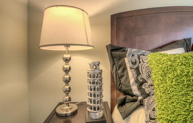 A lamp with a base resembling a tower is on a bedside table next to a bed with a green pillow.