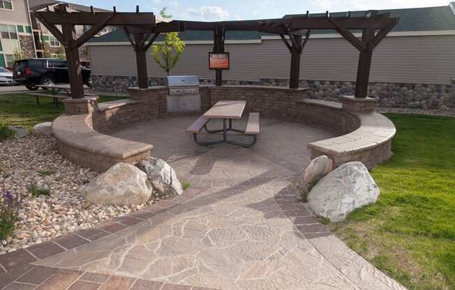 A welcoming outdoor picnic and grilling area here at Prairie Pines at the Ridge featuring a covered pergola with stone seating walls, a built-in stainless steel grill, and a central picnic table, surrounded by landscaped rock accents, green lawn, and paved walkways designed for relaxed community gatherings.