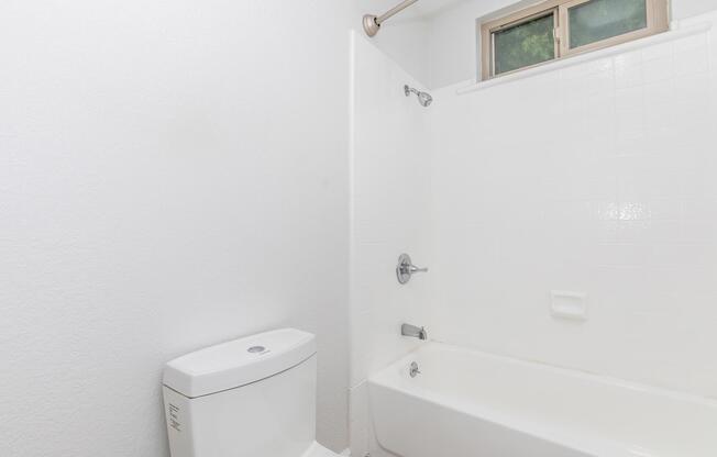 A clean, minimalist bathroom featuring a white bathtub and shower, a modern toilet, and a small window allowing natural light. The walls and tiles are also white, creating a bright and spacious feel. The overall design is simple and functional.