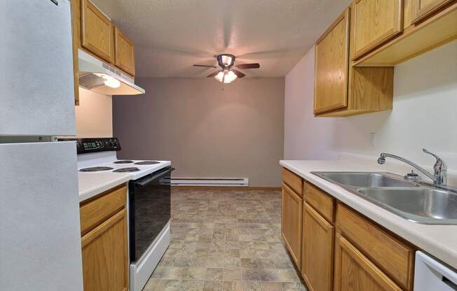 a kitchen with white appliances and a dining room in the background with a ceiling fan. Fargo, ND Dakota Manor Apartments